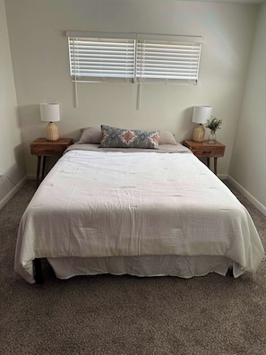 Un dormitorio con una cama grande, dos mesitas de noche, una ventana con persianas y suelo alfombrado.