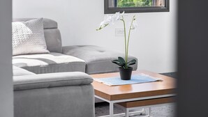 Una sala de estar con un sofá gris, una mesa de centro de madera con una planta en maceta y un ventanal con vistas a zonas verdes.