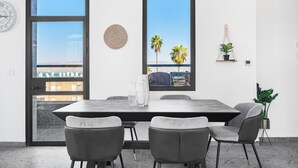 Un comedor moderno con una mesa grande y cuatro sillas, ventanas amplias con vistas a palmeras y un edificio, además de un reloj de pared.