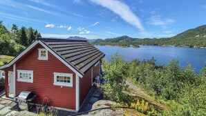 Une cabane rouge, dotée d’un toit en pente, surplombe un lac et est entourée de verdure et de collines.