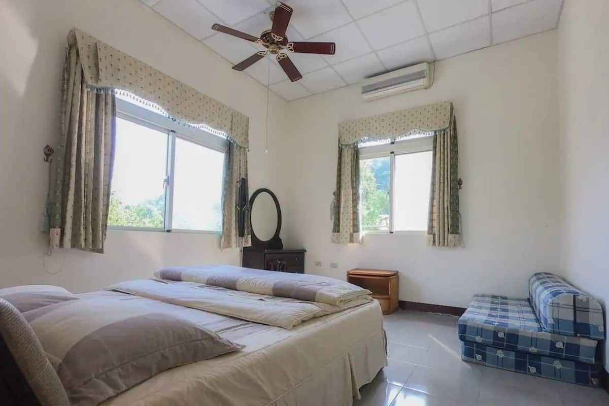 A bedroom with a bed, a ceiling fan, two windows with curtains, a small wooden table, and a blue sofa.