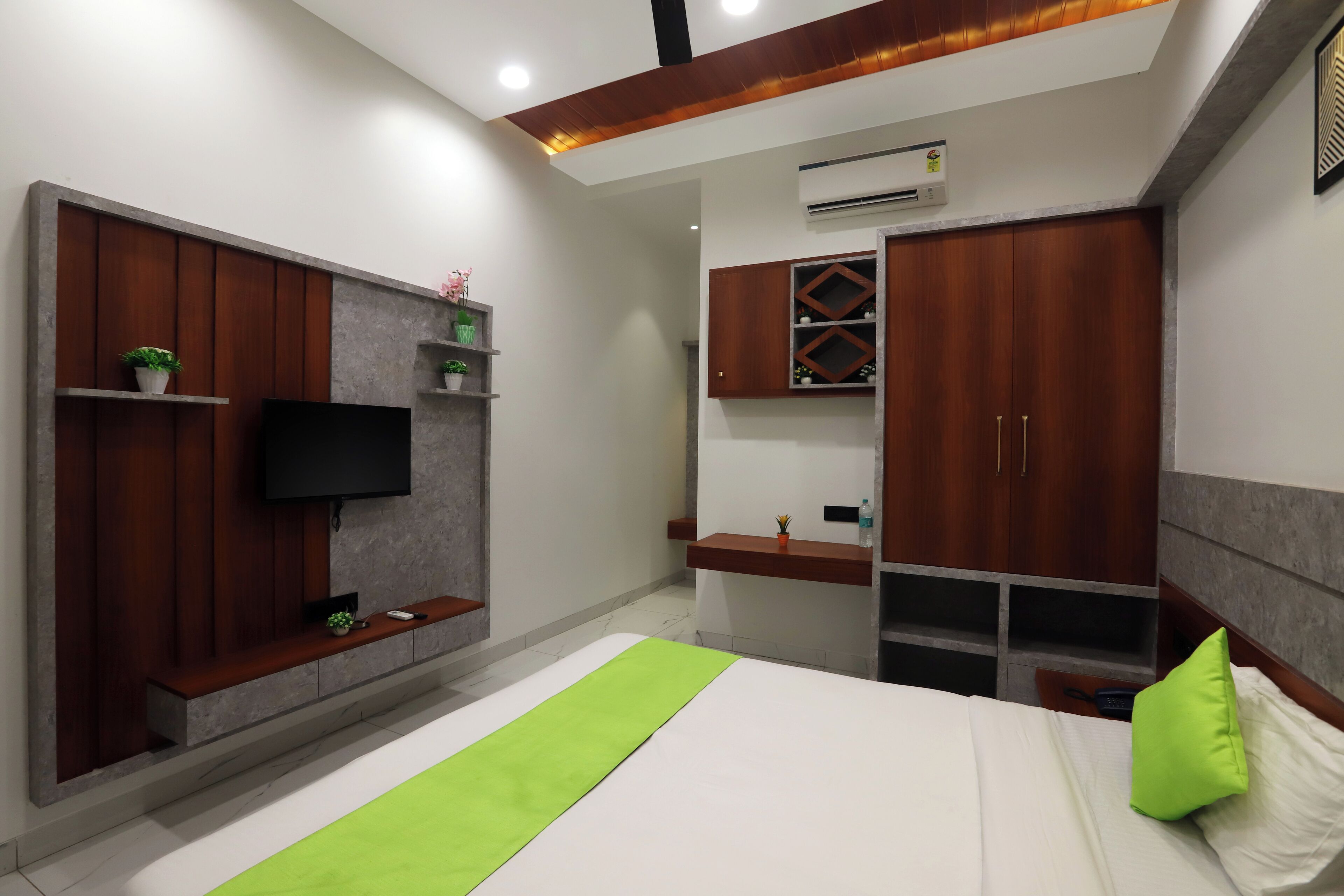 Un dormitorio moderno con cama, televisor montado en un panel de madera, armario y una pequeña estantería con plantas.