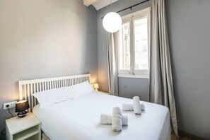 Cama bem arrumada com lençóis brancos e toalhas dobradas, luminária de cabeceira e janela com cortinas.
