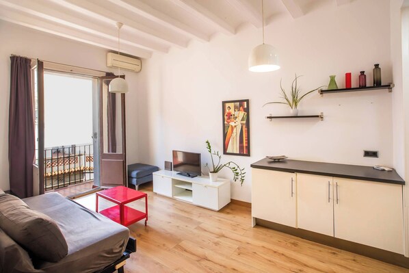 A modern living room with a sofa, a red table, a TV, and a kitchen area with cabinets and a countertop.