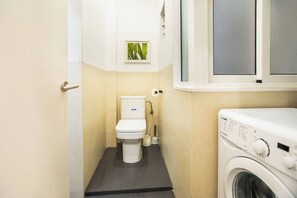Casa de banho compacta com sanita, máquina de lavar roupa e uma pequena janela com uma planta.