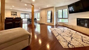 A spacious living room with hardwood floors, a large TV mounted on the wall, and a fireplace. - Whispering Pines: Garage Parking & Game Room (Pollock Pines)