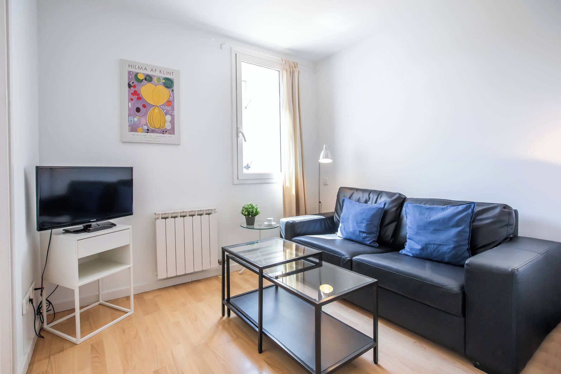 A modern living room with a flat-screen TV, a glass coffee table, a black leather sofa with blue cushions, a radiator, and a window with curtains.