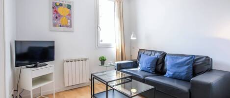 A modern living room with a flat-screen TV, a glass coffee table, a black leather sofa with blue cushions, a radiator, and a window with curtains.