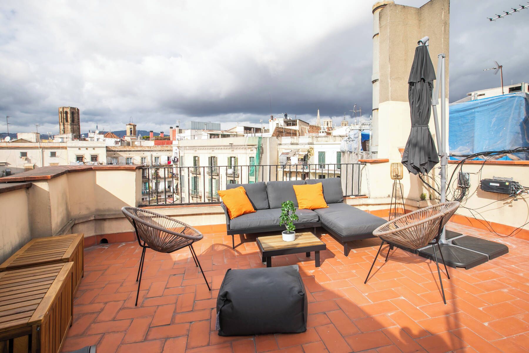 A rooftop terrace with a grey sofa, two wicker chairs, a small table, and a black ottoman.