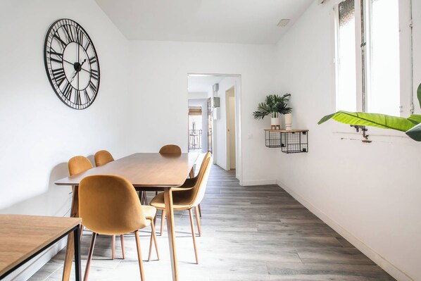 Un coin repas avec une table et des chaises en bois, une grande horloge murale et une plante en pot près d’une fenêtre.