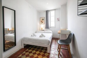 Une chambre à coucher comprenant un lit, un bureau, une chaise, un miroir, une fenêtre avec des rideaux et une étagère fixée au mur.