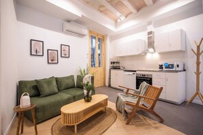 Un espace de vie compact comprenant un canapé vert, une table basse en bois, une chaise en bois et un coin cuisine avec des meubles blancs et des appareils électroménagers.