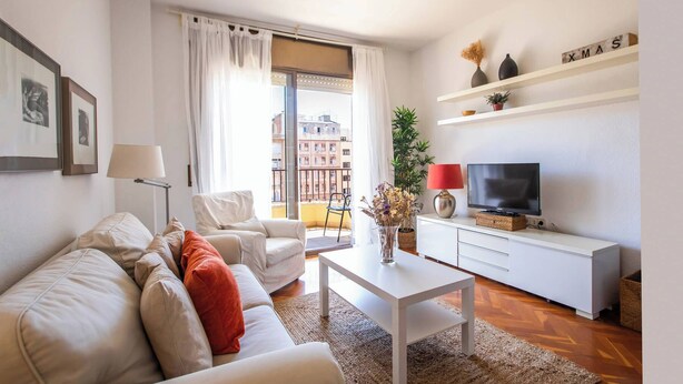 Ein modernes Wohnzimmer mit Sofa, Sesseln, Couchtisch, Fernseher und einem Balkon mit Blick auf die Stadt.
