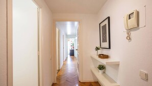 Ein Korridor mit Holzboden, weißen Wänden und einer Tür am Ende. Regale mit Dekorationen und ein gerahmtes Bild sind an der Wand angebracht.
