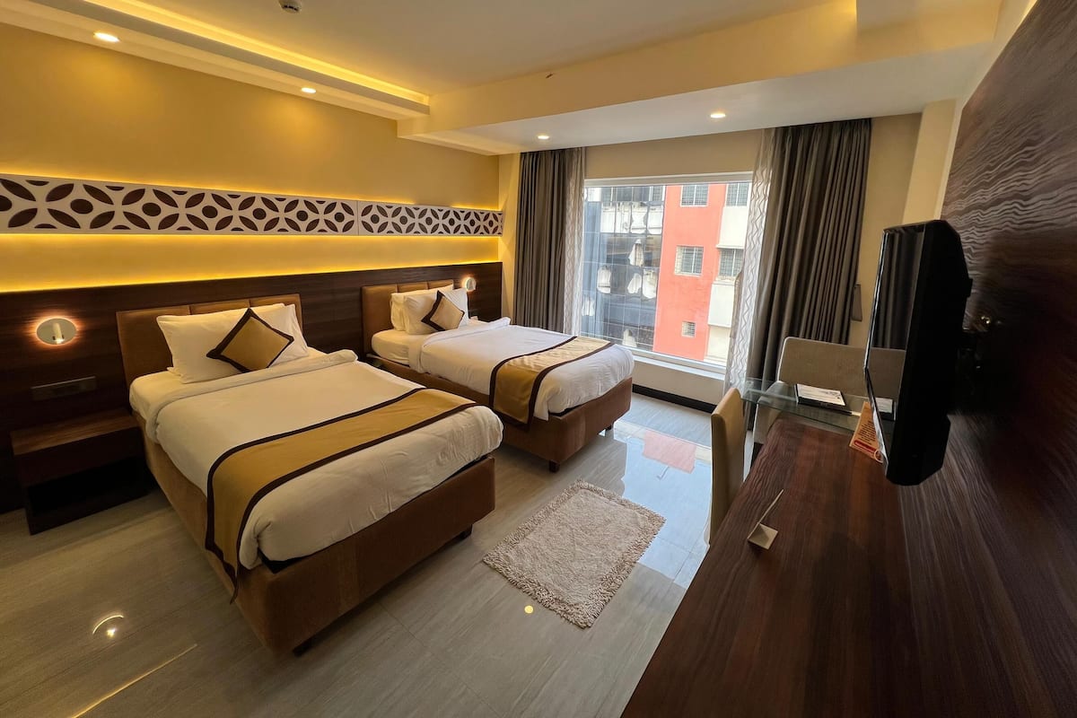 A hotel room with two beds, a TV, a desk, and a view of a building.