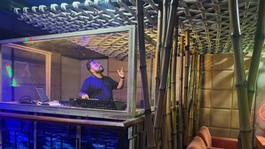 Un equipo de DJ con una computadora portátil y equipo de audio, rodeado de estructuras de bambú y una zona de estar con sillas naranjas.