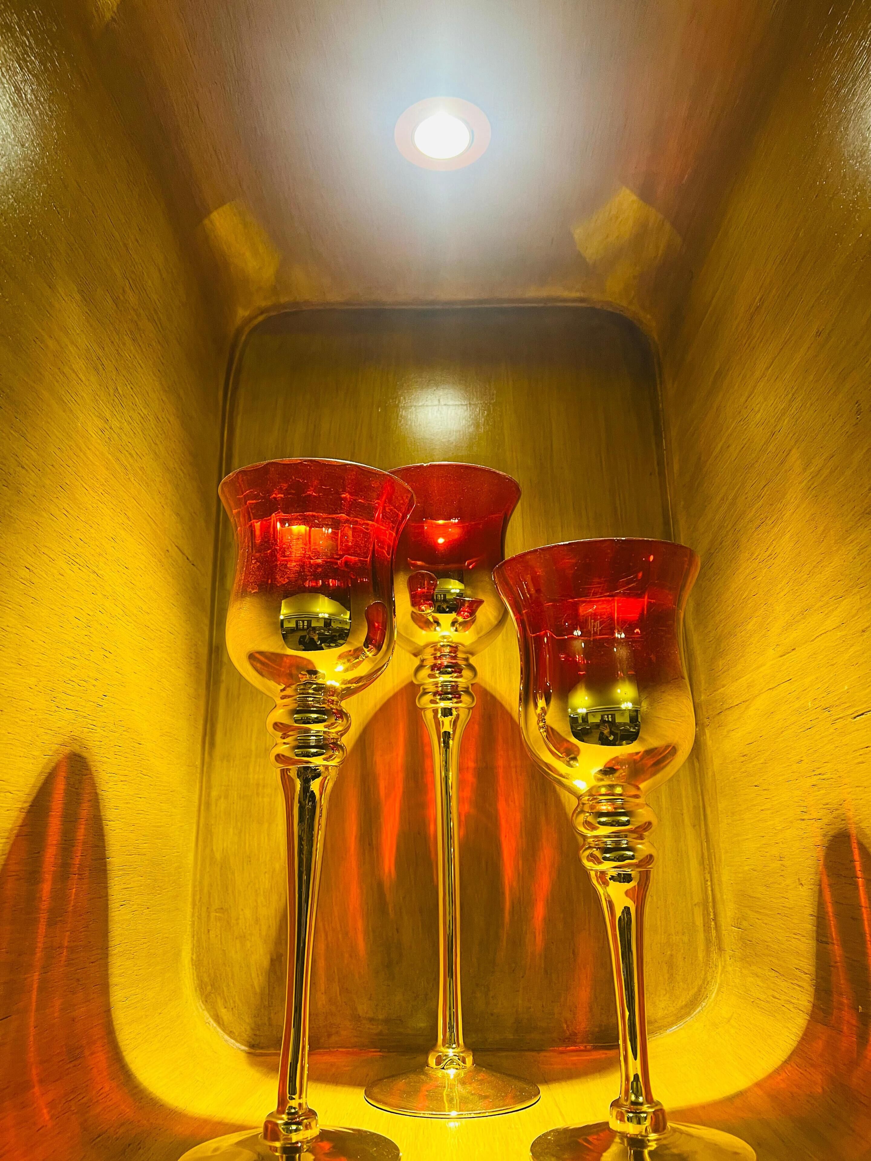 Tres copas de vino con tallos rojos y base transparente, colocadas dentro de un recipiente metálico reflectante.