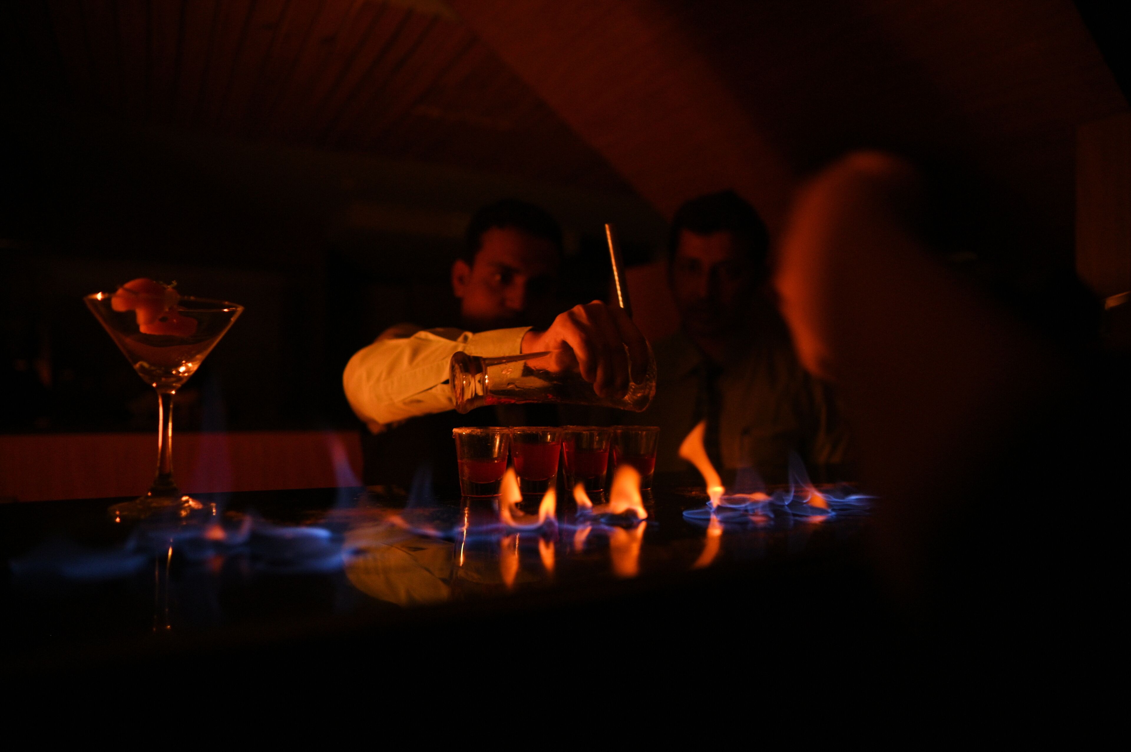 A dimly lit bar area with a flaming drink, a martini glass, and a person pouring a drink.