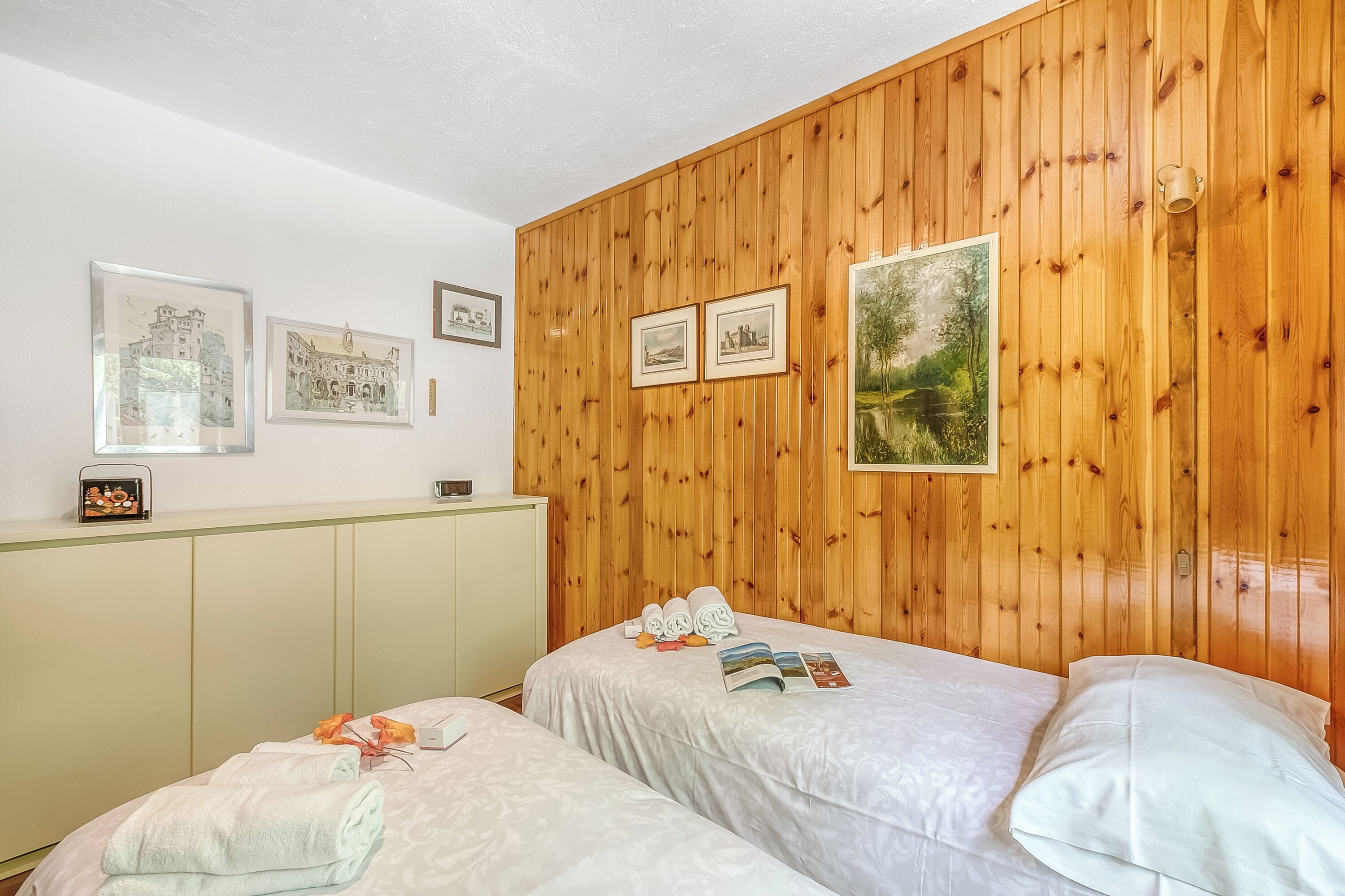 A room with two beds, wooden panel walls, and framed pictures.