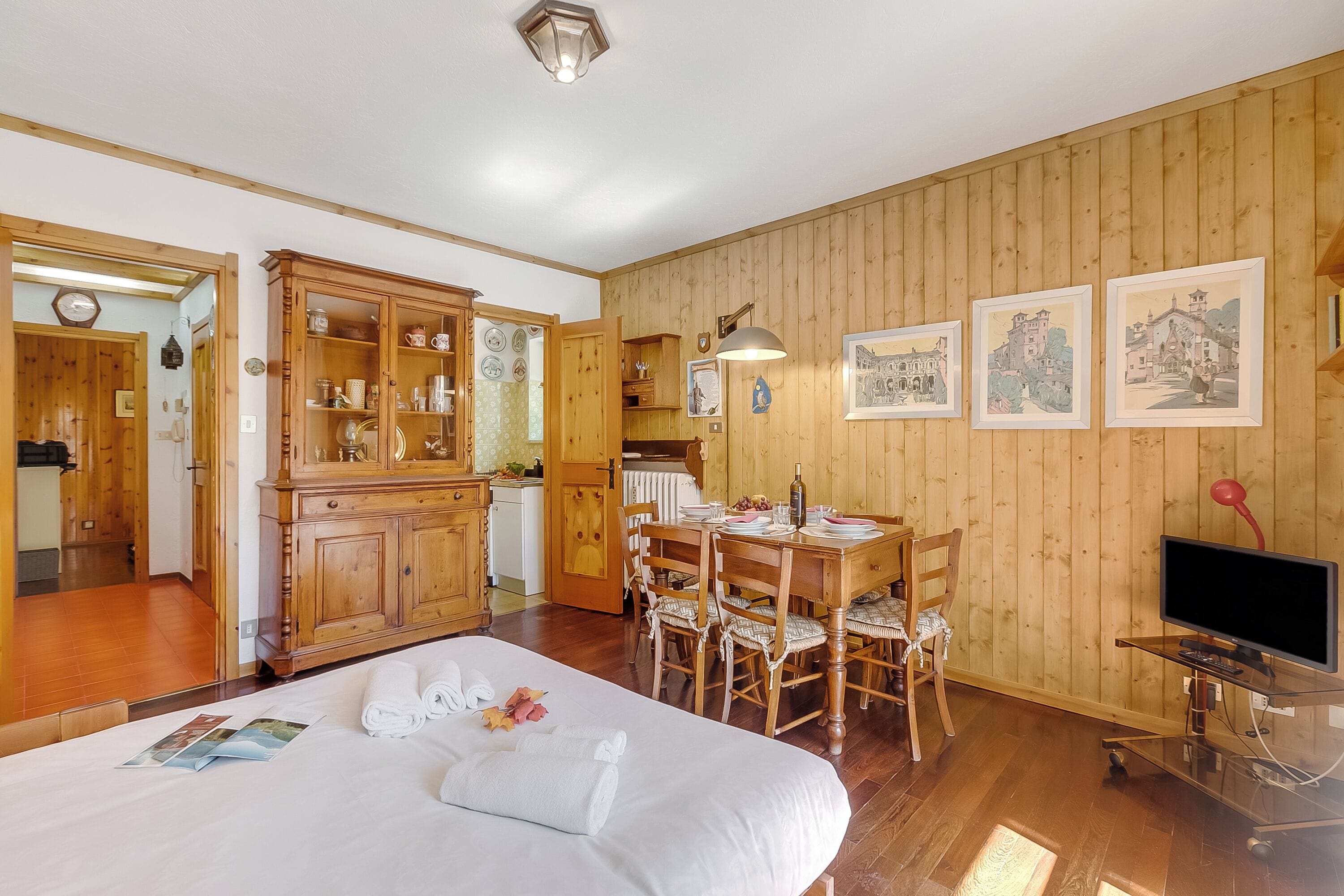 Una habitación con paneles de madera, una cama, una mesa de comedor, sillas, un televisor y una zona de cocina.
