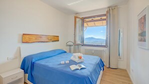 A bedroom with a bed, a nightstand, a window with a view, and a painting on the wall. - Il Vecchio Faggio: Lake Maggiore View Terrace (Gignese)