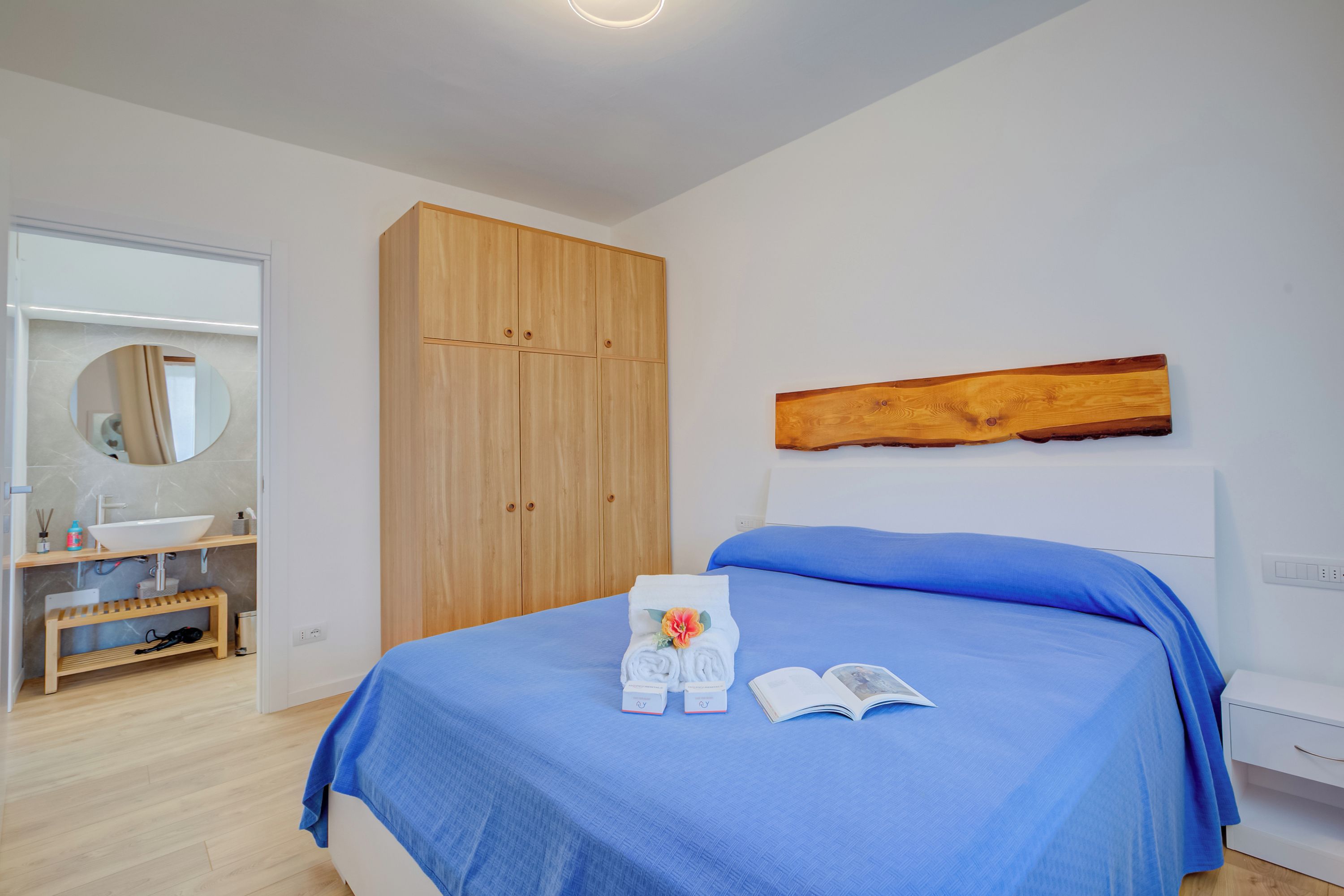 A bedroom with a bed, a wooden wardrobe, a mirror, and a bathroom with a sink and vanity.