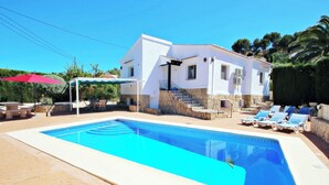 Une piscine rectangulaire bordée de pierres, un bâtiment blanc avec une base en pierre et un parasol rouge.