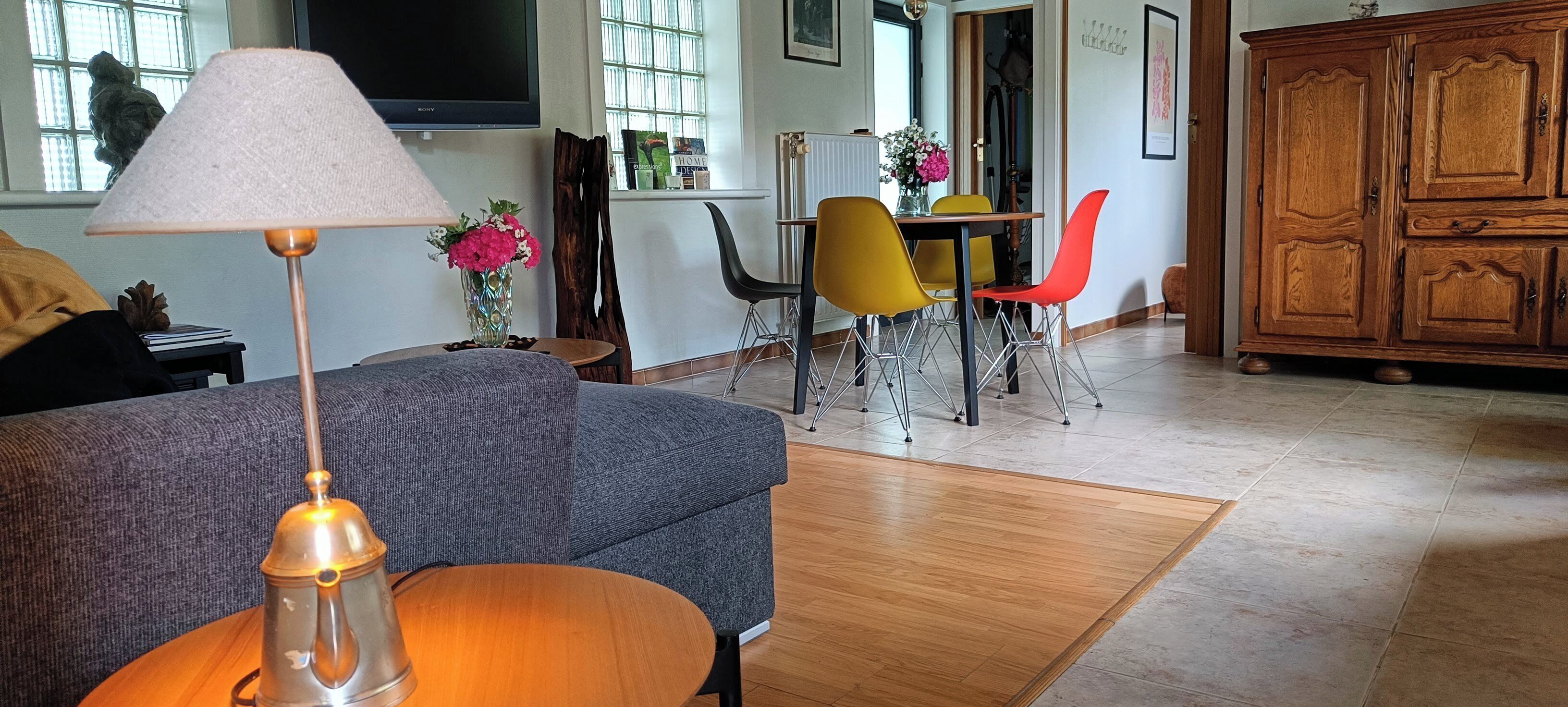 Een woonkamer met een grijze bank, een houten salontafel, een lamp, een televisie, een eettafel met gele en rode stoelen, en een houten kast.