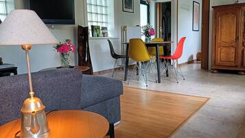 Een woonkamer met een grijze bank, een houten salontafel, een lamp, een televisie, een eettafel met gele en rode stoelen, en een houten kast.