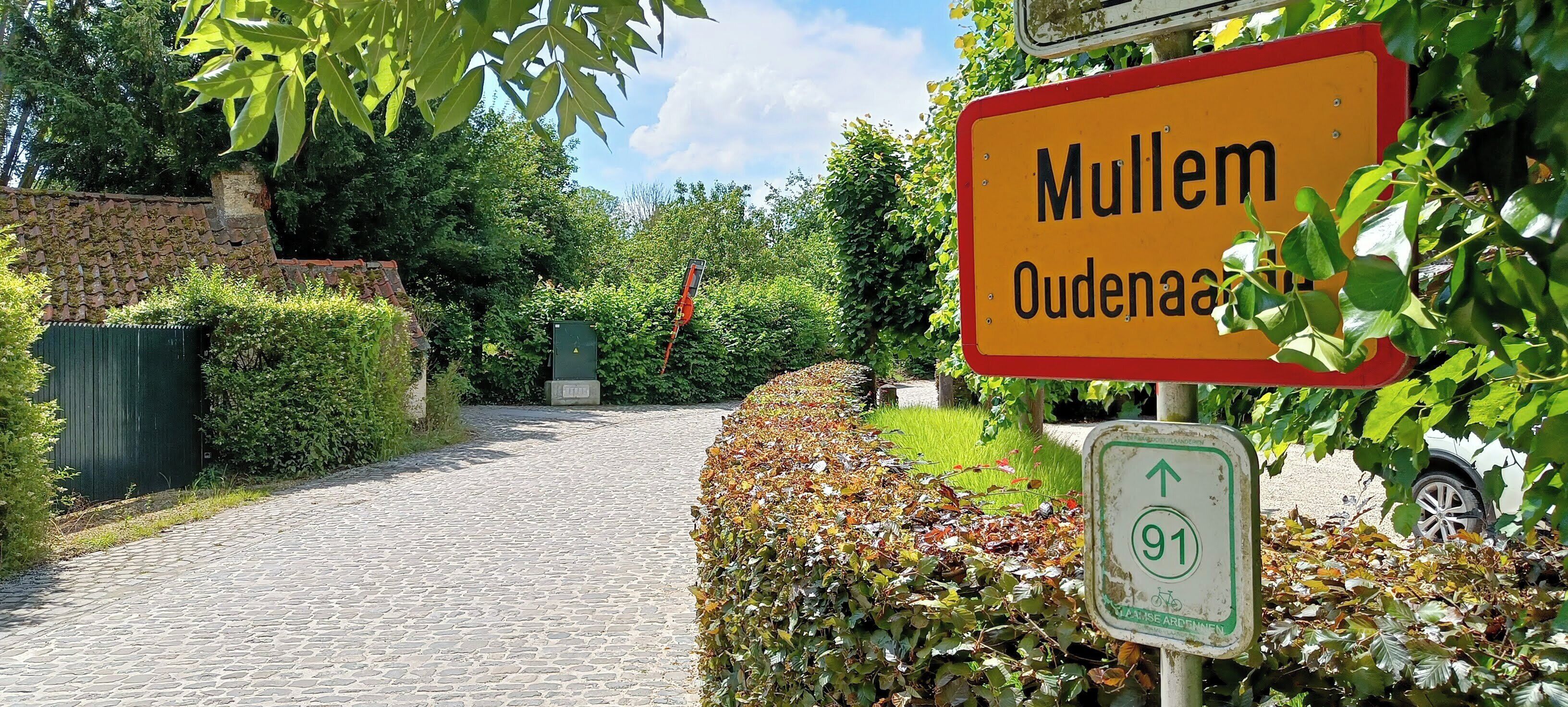 Ein Kopfsteinpflasterweg führt zu einem Gebäude, an dem die Schrifttafel "Mullem Oudenaar" angebracht ist.