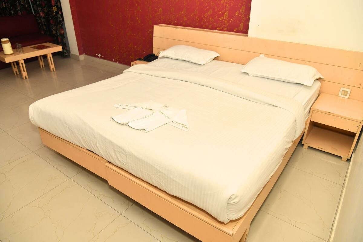 Una cama bien hecha con ropa de cama y toallas blancas, una mesita de noche de madera con una lámpara, y una pared roja con papel tapiz floral.