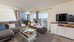 A modern living room with a sofa, coffee table, and TV. There are large windows with curtains, a view of the sea, and a balcony.