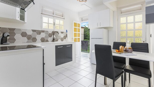 A modern kitchen with white cabinets, a black countertop, and a hexagonal tile backsplash. There is a dining area with a white table and black chairs.