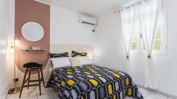 Een slaapkamer met een bed, een ronde spiegel, een kruk, een airconditioning en een raam met gordijnen.