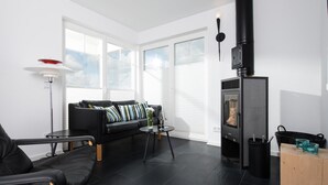 A modern living room with a black leather sofa, a small round table with a vase and wine glass, a black wood-burning stove, and large windows with blinds. - Cozy home in Olpenitz with sauna (Olpenitz)