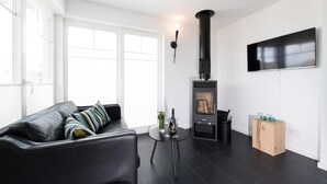 A modern living room with a black leather sofa, a round black table with a bottle and glasses, a black wood-burning stove, and a mounted flat-screen TV. - Cozy home in Olpenitz with sauna (Olpenitz)