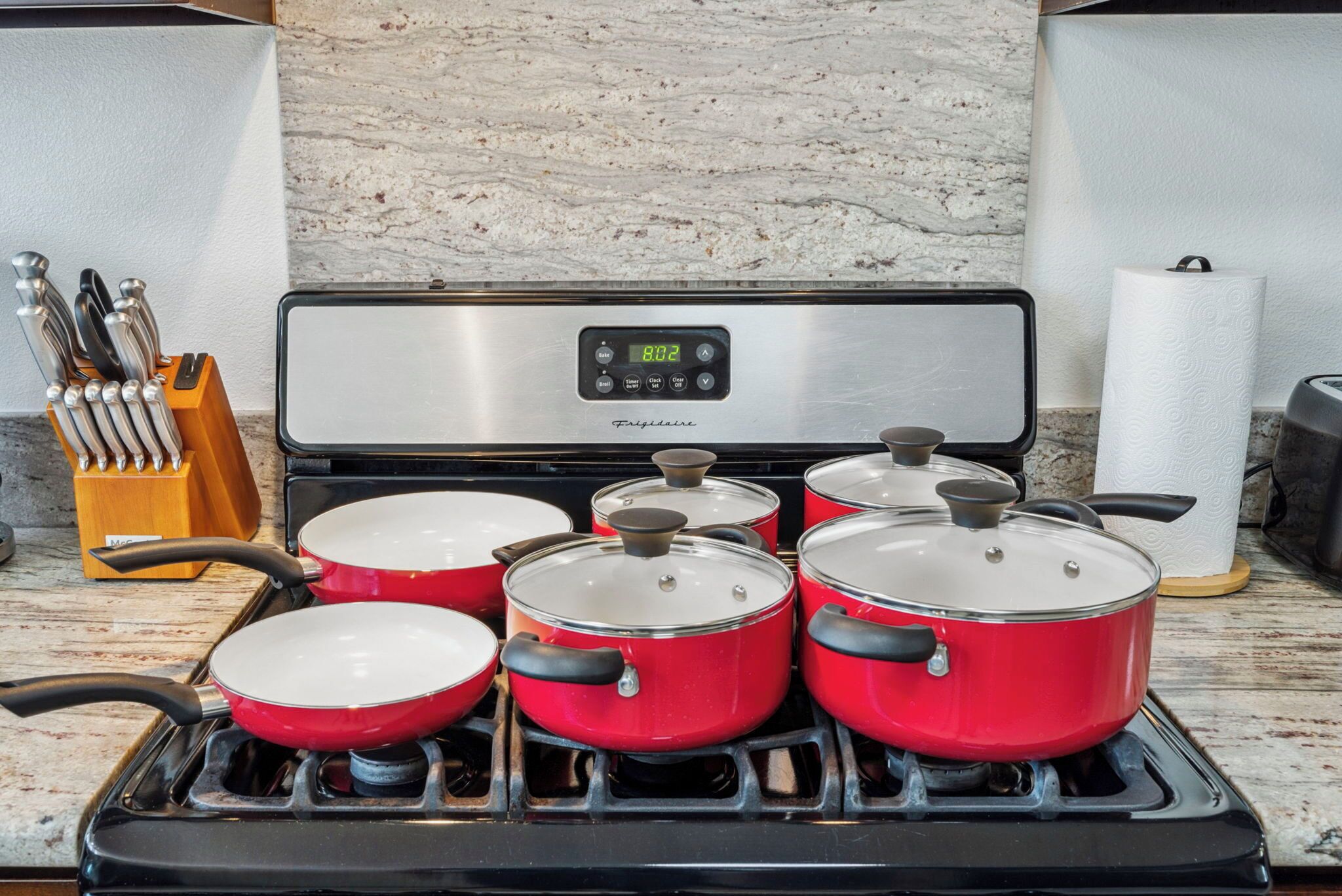 A kitchen with a stainless steel stove, six red pots, a knife block, and a marble countertop.