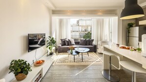 Un salon moderne avec un canapé, une table basse et des tabourets de bar. Un téléviseur est fixé au mur, et il y a une fenêtre avec des rideaux.