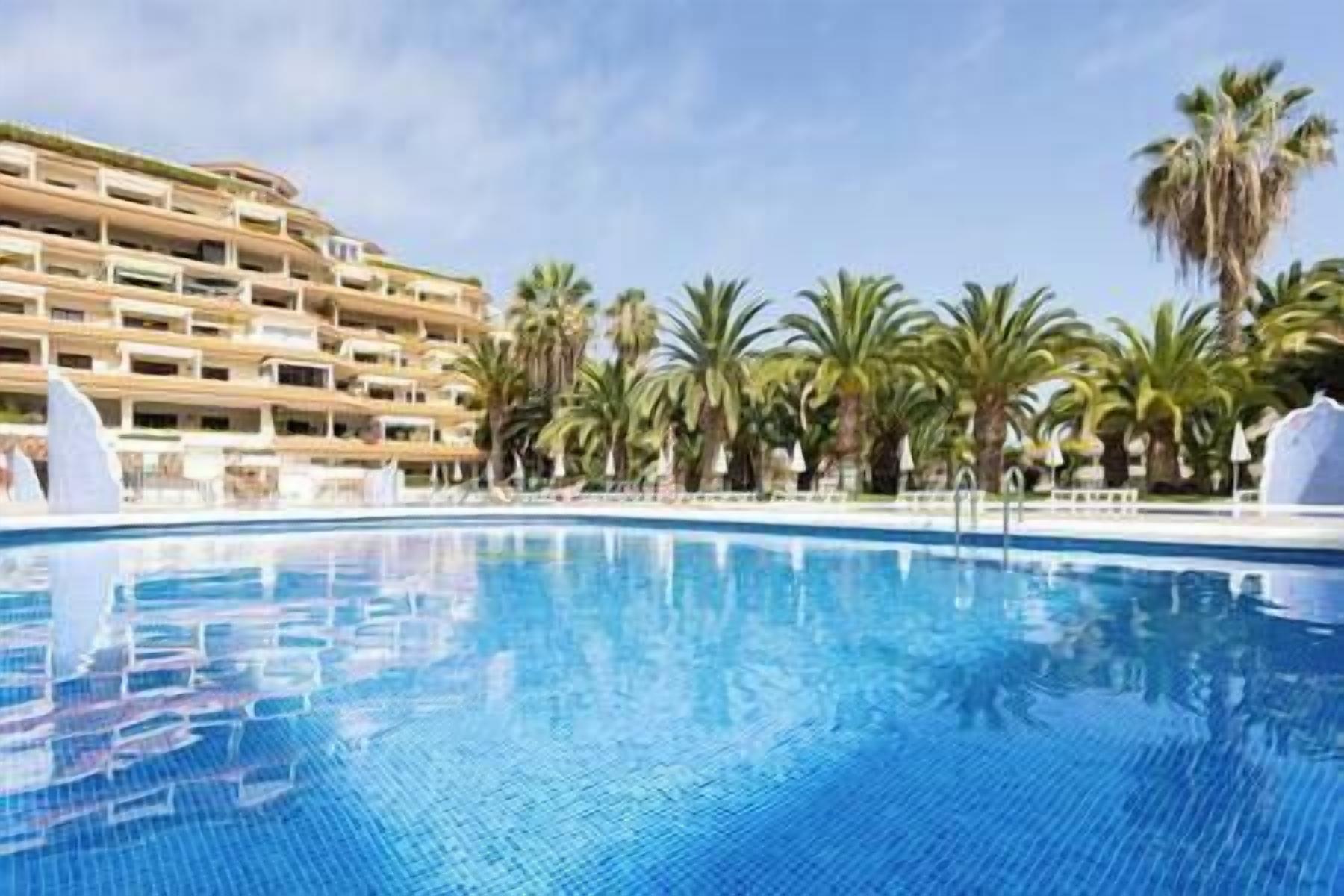 A swimming pool with a clear blue water surface, surrounded by palm trees and a multi-story hotel building with balconies.