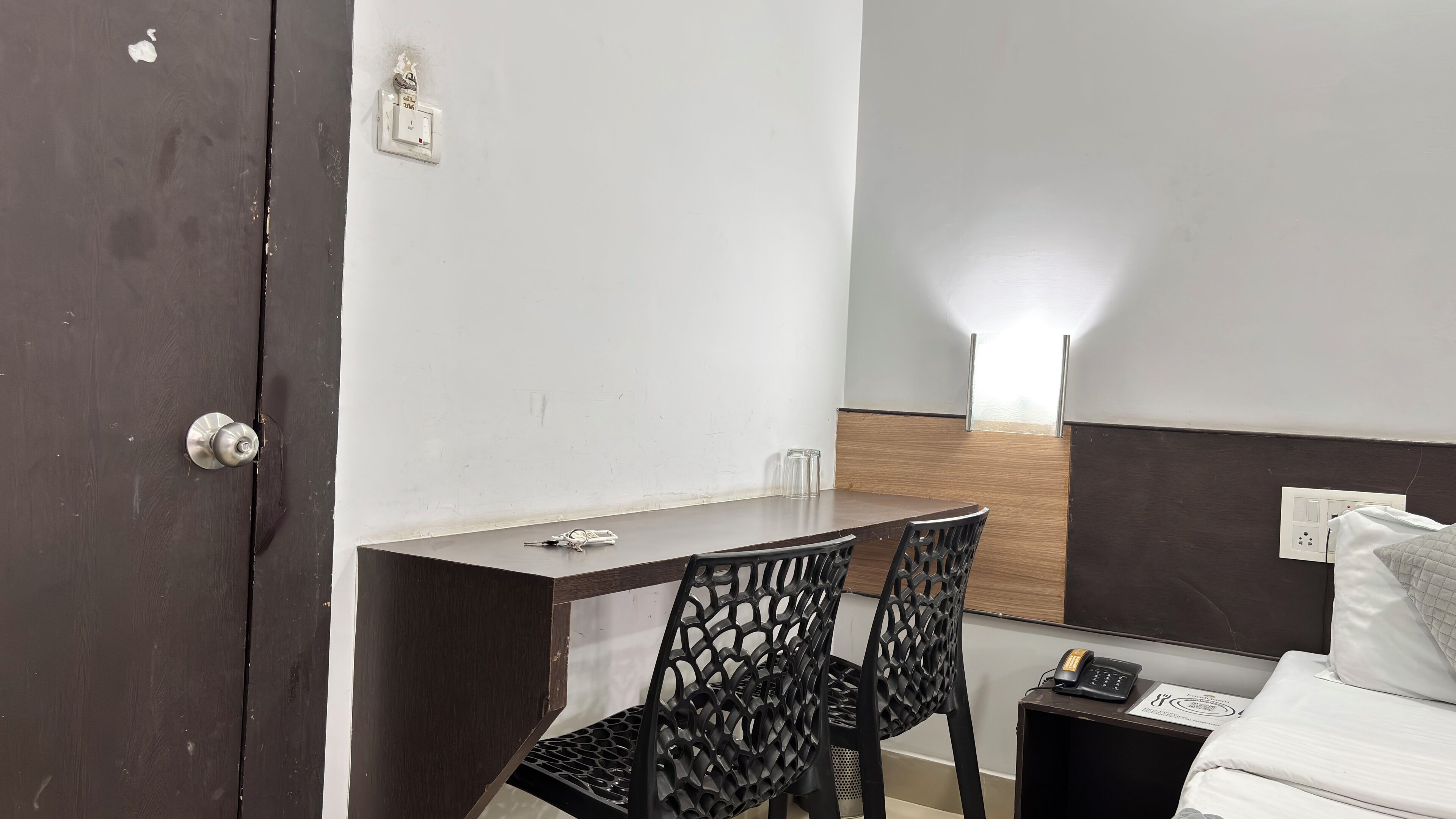 Habitación de hotel con una puerta de madera, una pequeña mesa con sillas, una mesita de noche con un teléfono y una lámpara fijada a la pared.