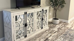 Un meuble blanc sur lequel est posée une télévision, un pot de fleurs et un tapis à motifs.