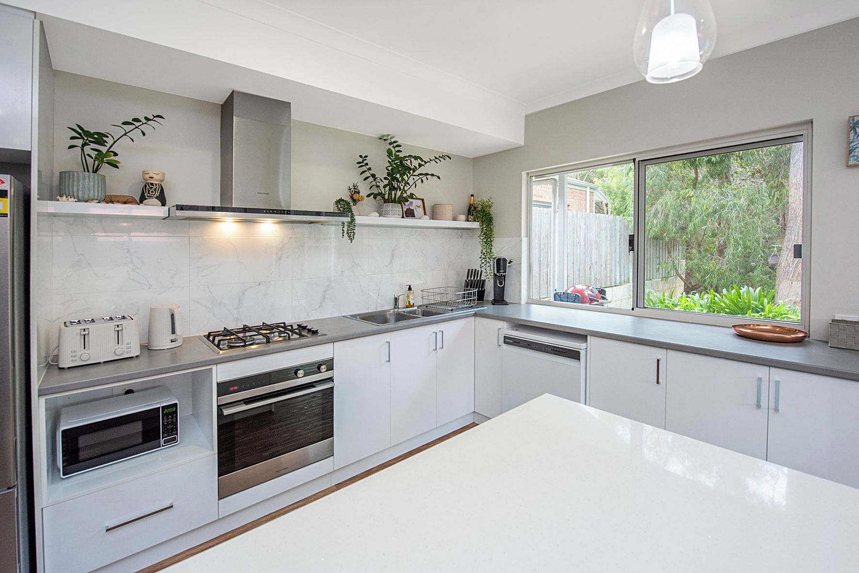 Una cocina moderna con gabinetes blancos, estufa a gas, microondas y lavavajillas. Se ve un ventanal con vistas a la vegetación.