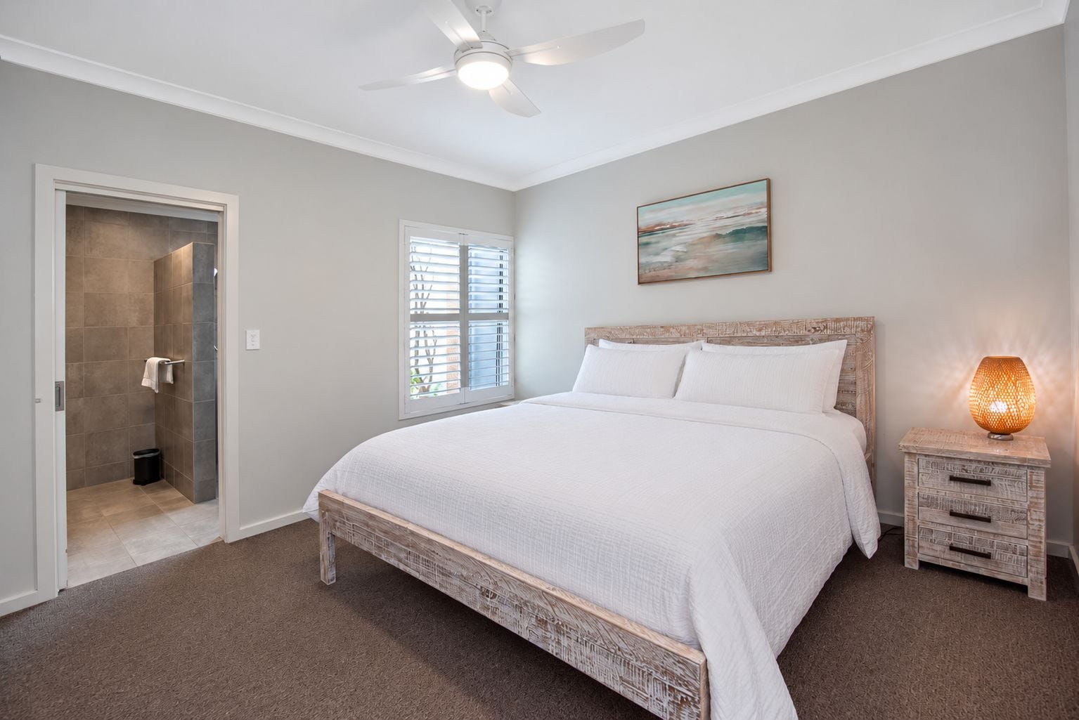 A bedroom with a bed, a nightstand, a wall-mounted lamp, a ceiling fan, a window with blinds, and a door leading to a bathroom.