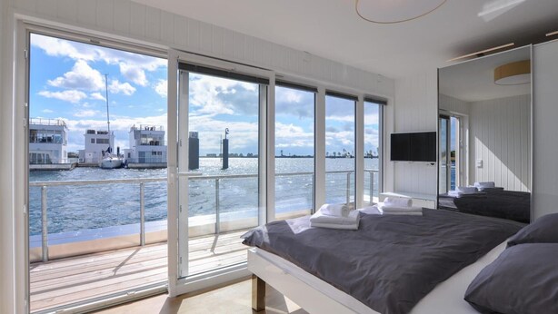 A modern bedroom with a large bed, a TV, and a view of the water and boats. - Beach front ship in Olpenitz with sauna (Olpenitz)
