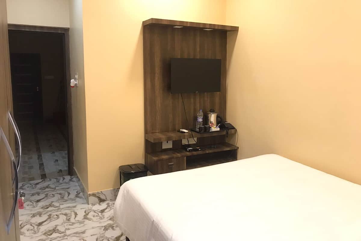 A hotel room with a bed, a flat-screen TV mounted on a wooden panel, a small desk with a phone and a water bottle, and a trash bin.