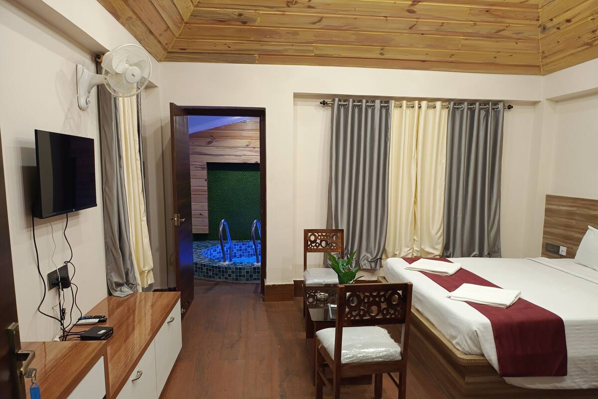 A hotel room with a wooden ceiling, a TV mounted on the wall, a bed with a red blanket, a desk with a laptop, and a small table with a chair.