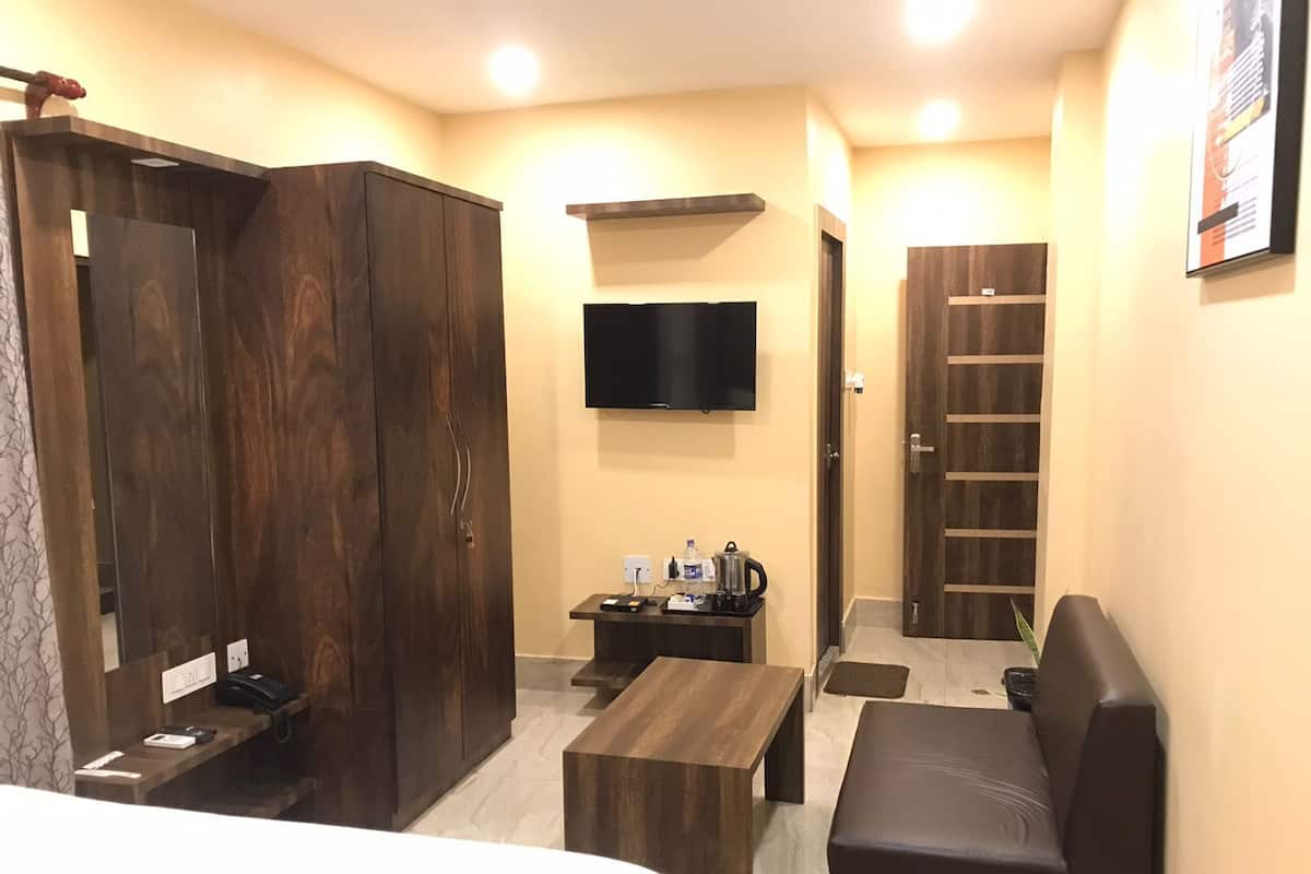 A hotel room with a bed, a wooden wardrobe, a small table with a kettle, a flat-screen TV, a sofa, and a door with a window.