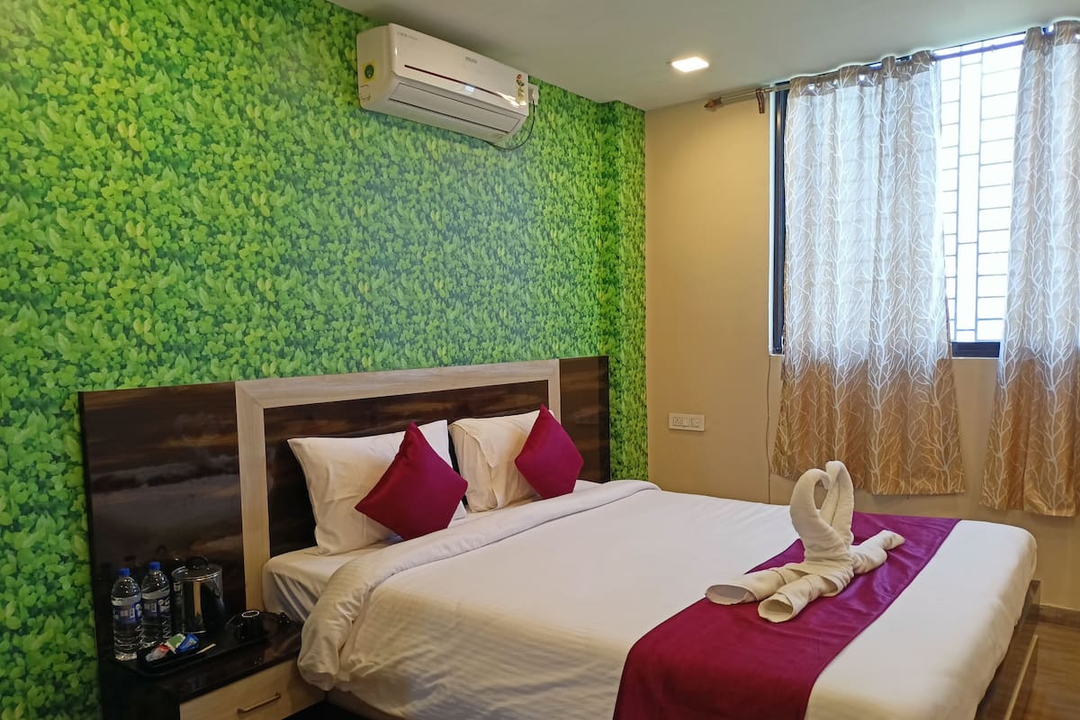 A hotel room with a green leaf-patterned wall, a wooden headboard, white bedding, red pillows, a tray with toiletries, and a window with patterned curtains.