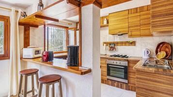 A compact kitchen with wooden cabinets, a microwave, a stove, and a sink. There are two stools and a window providing natural light.