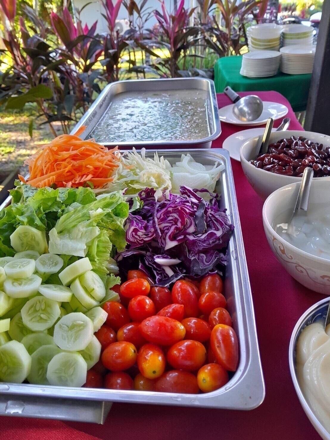 A buffet table with various food items including sliced cucumbers, cherry tomatoes, shredded carrots, and mixed greens.